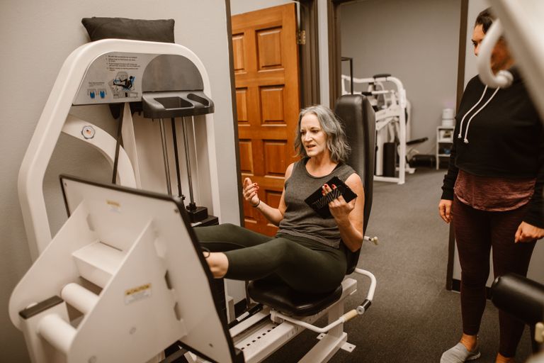 Senior woman working out on a leg press machine with a trainer at a gym.