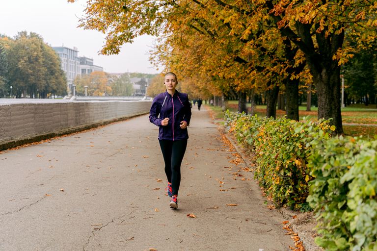Woman jogging on a tree-lined path in the park with autumn leaves.
